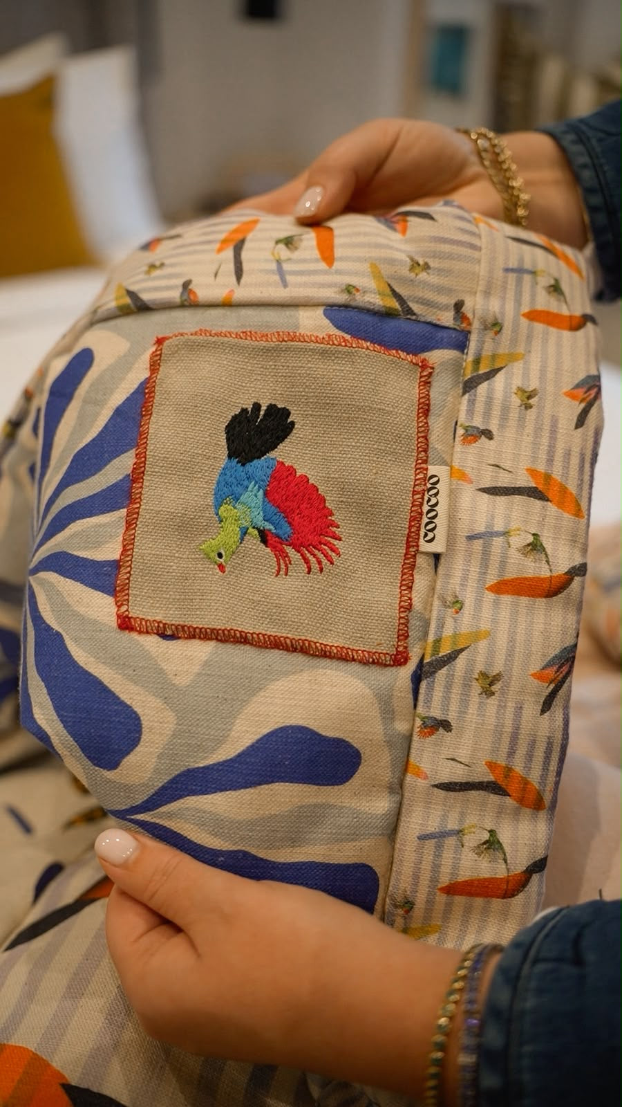 A close-up of an embroidered Loerie bird on patterned fabric being held by hands. The bag features a blue and beige abstract pattern on one side, a striped side with orange bird motifs, and a square embroidered patch showing a colorful Loerie, featuring the new quilts by The Coocoo Club.