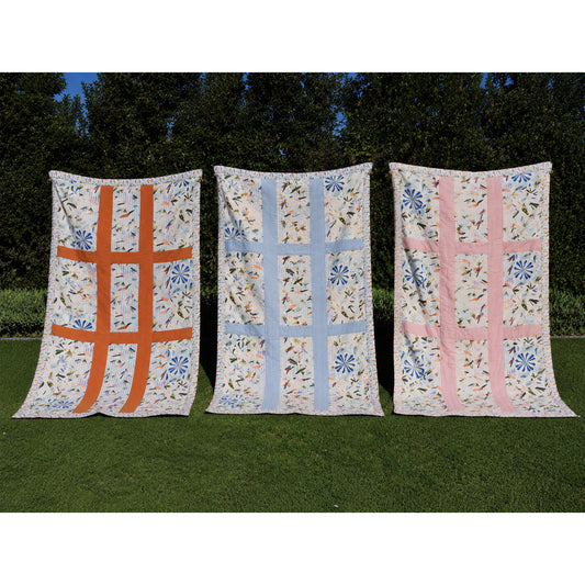 Three vintage style quilts with floral patterns are displayed side-by-side on a green lawn against a backdrop of dark green trees. The left quilt features an orange grid pattern on a white floral background, the middle quilt has a light blue grid pattern, and the right quilt has a pink grid pattern, all designed by The Coocoo Club.